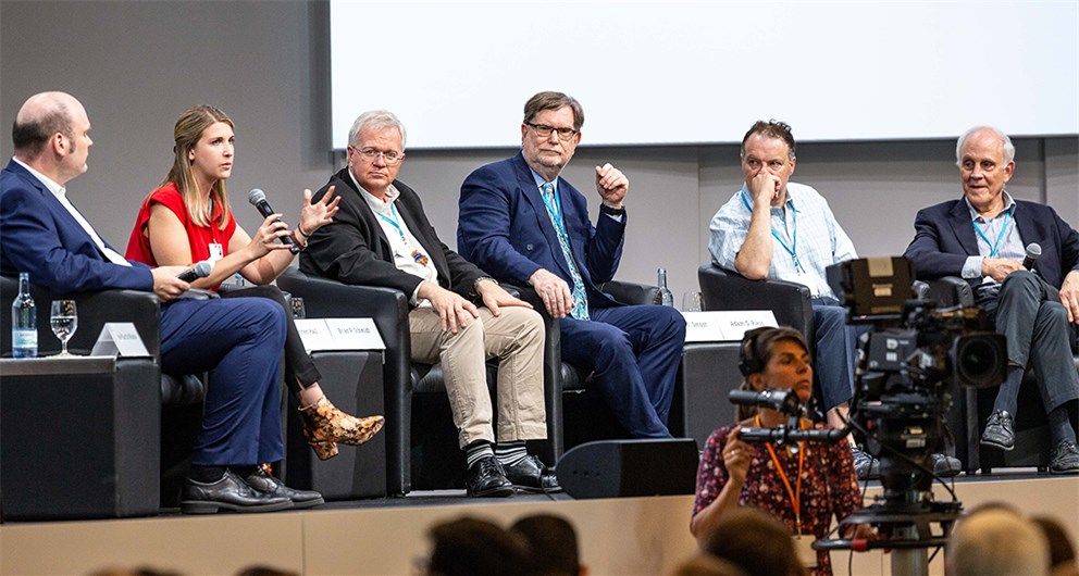 A panel of speakers sits on a stage, there is one blond white woman surrounded by older white men.