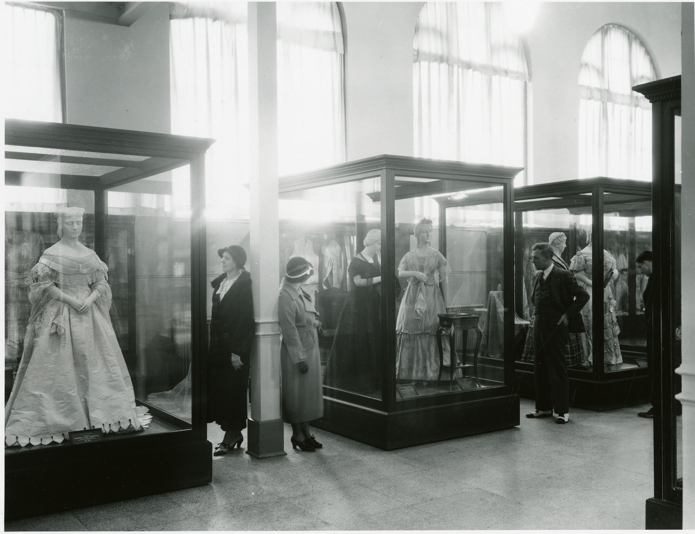 A large room with museum cases holding mannequins wearing gowns. A few people look into the cases.