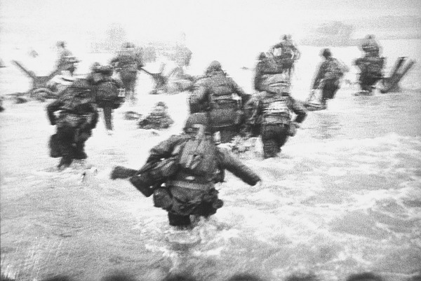 A grainy black and white image of troops walking through ocean water.