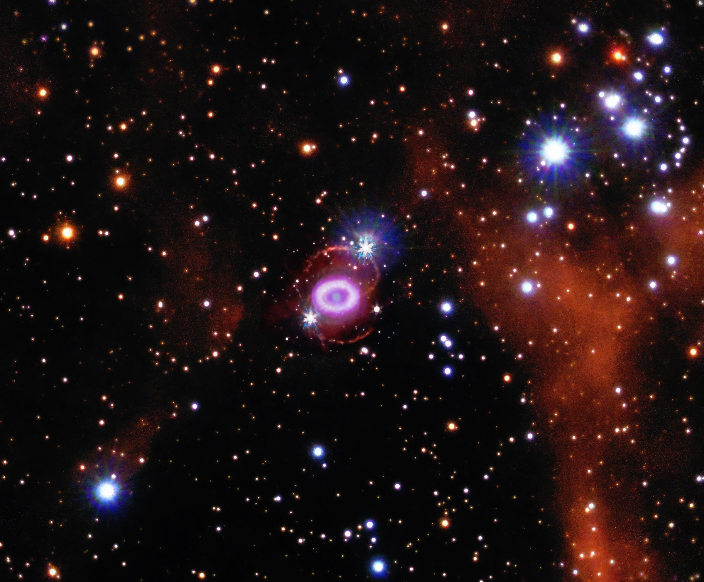 At the center of this composite image is a small object resembling a glowing pink Cheerio. Inside this ring is a pale, steel blue dot containing debris from the star that exploded.. The ring sits at the center of a ghostly figure 8, outlined in brick orange. This entire structure is surrounded by a packed field of stars, specks and dots in white, blue, and orange. A long, brick orange cloud hovers near the left edge of the image.