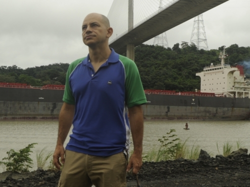 Carlos Jaramillo stands in front of a bridge over the Panama Canal on a cloudy day.