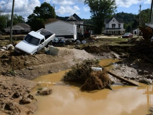 A big pool of muddy water surrounded by small homes and a white pickup truck that's angled into a ditch.