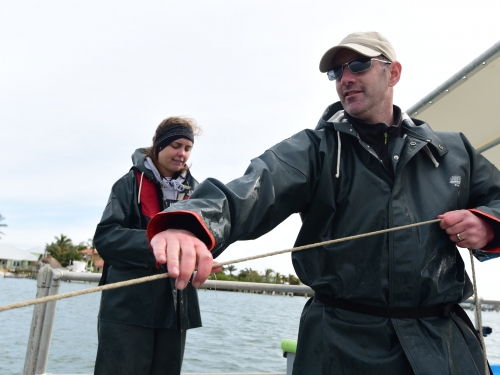 : A man in a black jacket with sunglasses and a young woman stand on a boat. The man holds a line of rope in front of him.