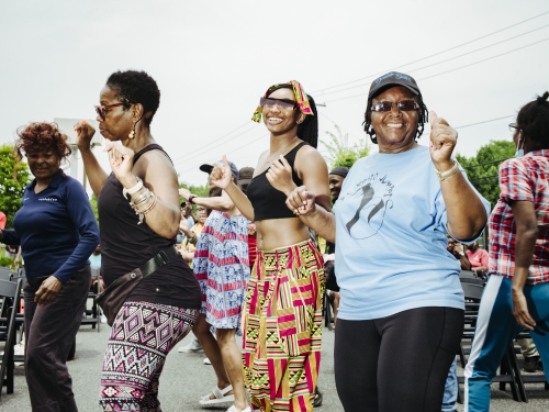 Three women in colorful clothing dance with their arms raised amongst a light crowd.