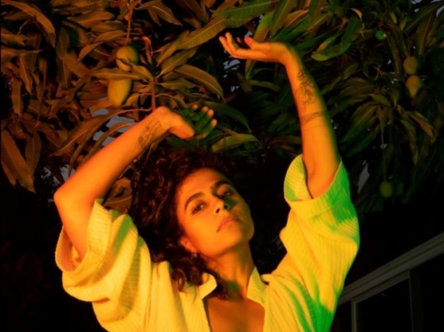 Woman with elbow-length sleeves holds her arms up above her head, with greenery in background.