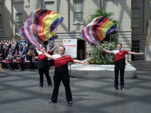 Three young women stand in a triangle formation, holding large flags with rainbow colors.