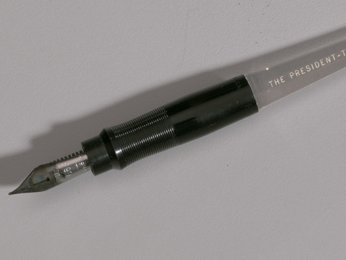 A pen with a black plastic grip and a clear plastic body, with [THE PRESIDENT- THE WHITE HOUSE] printed in white ink.