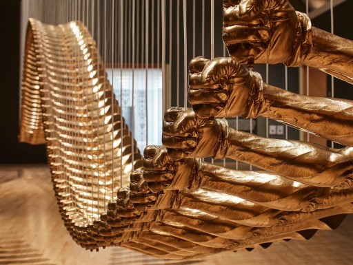 A series of bronze hands with gloves on them hang in suspension at different length, creating a wave effect.