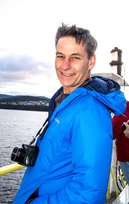 Man with short grayish-black hair smiles, standing on a boat with yellow handrail and wearing blue puffy jacket.