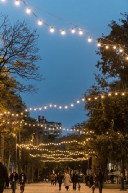 Photograph of a brick street, lined with trees, people walking, and string lights in a zig-zag pattern.