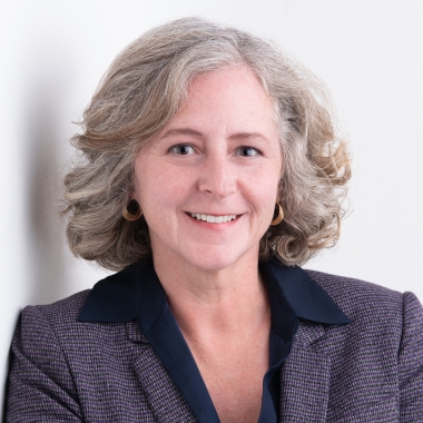 Woman with shoulder-length, silver hair stands against white wall, smiling.